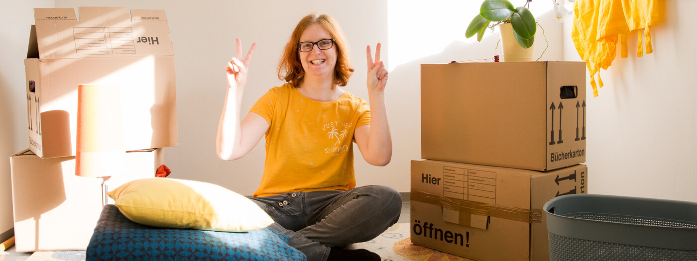 Junge Frau sitzt erfreut zwischen Kartons in ihrer eigenen Wohnung